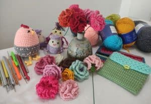 Craft desk with colorful crochet items: flowers, small chick, yarn skeins, crochet hooks, and a green turquoise crochet pouch on a white table.
